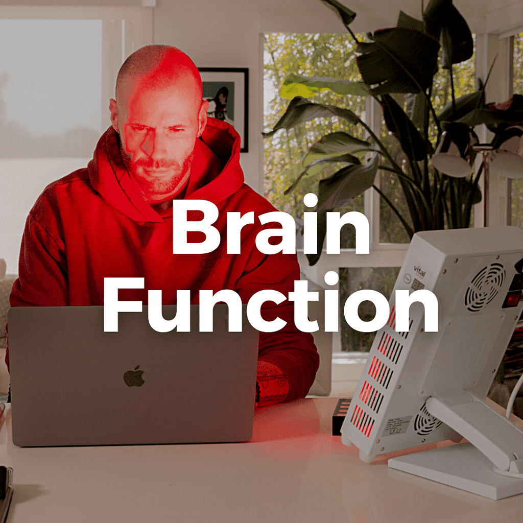 A person in a red hoodie is focused on their laptop with "Brain Function" text, while benefiting from red light therapy in a well-lit room.