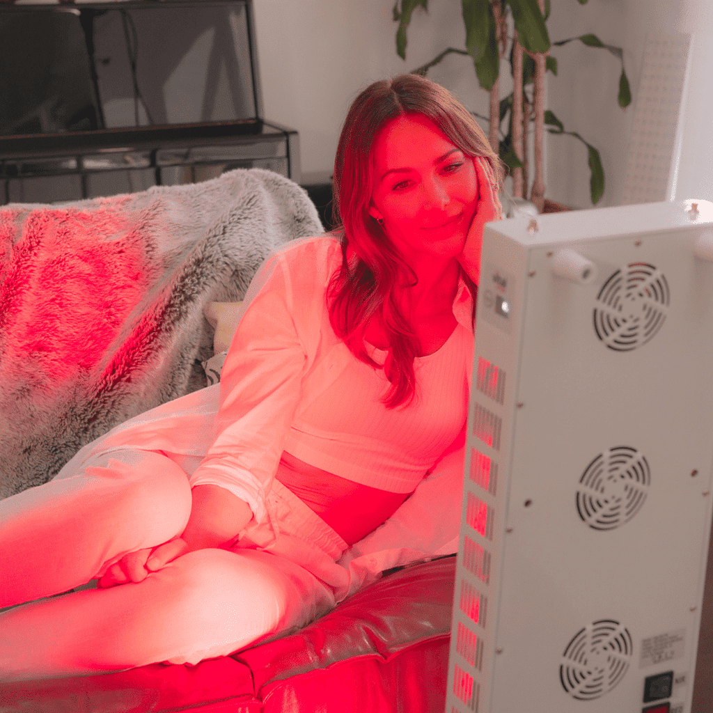 A woman in white clothing enjoys a calming moment on a sofa, bathed in the soothing glow of red light therapy from a nearby device.