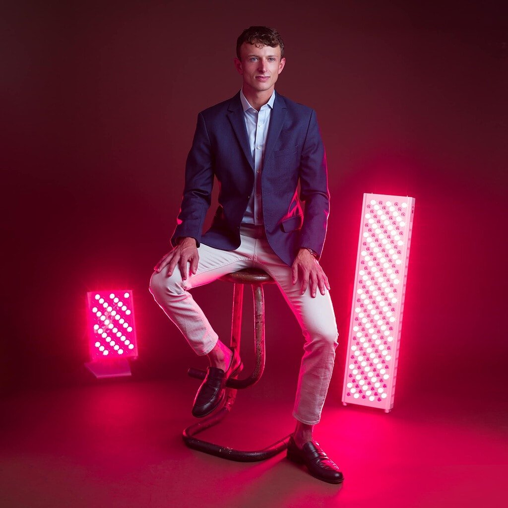 A man in a navy blazer and white pants sits on a stool between two vertical pink LED light panels, evocative of red light therapy benefits, against a dark background.