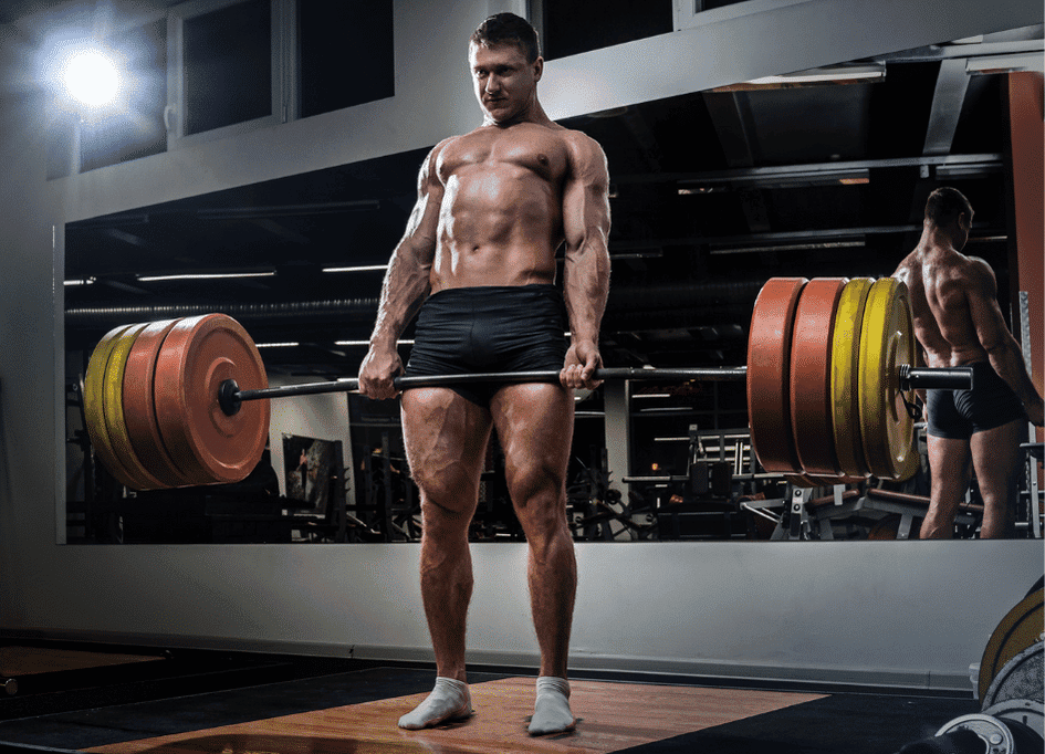 A muscular person performs a deadlift with a heavily loaded barbell in a gym, standing on a wooden platform. A large mirror and gym equipment are visible in the background, highlighting the dedication put into their fitness journey—perhaps boosted by the benefits of red light therapy for recovery.