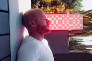 A man in a white shirt leans against a wall under a bright red light panel, eyes closed, as if assessing his next move after checking the wholesale inventory dashboard, outdoors near plants.