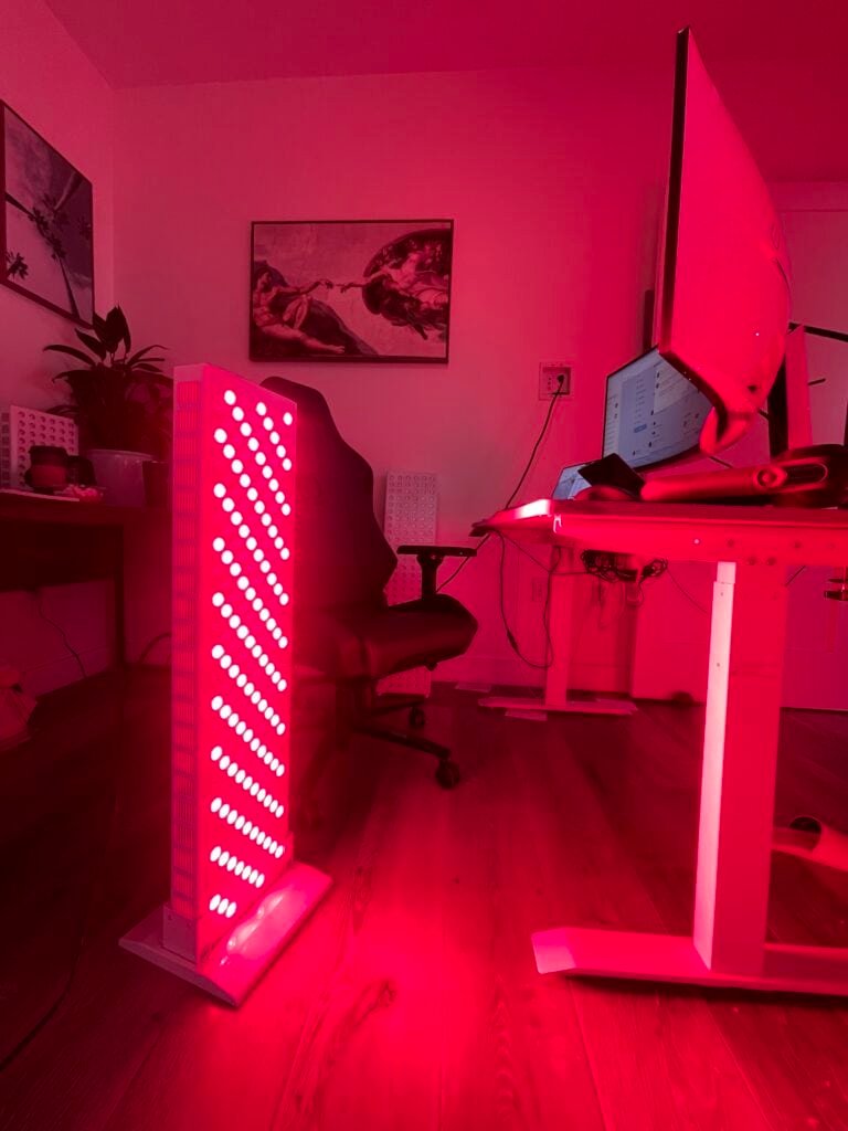 A room illuminated by red light from a tall, rectangular lamp beside a desk with a computer. A chair and wall art are visible in the background.