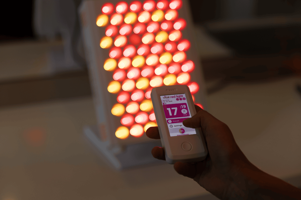 A hand holds a remote displaying settings for the Vital Pro 2.0 red light therapy device, which is illuminated in the background.