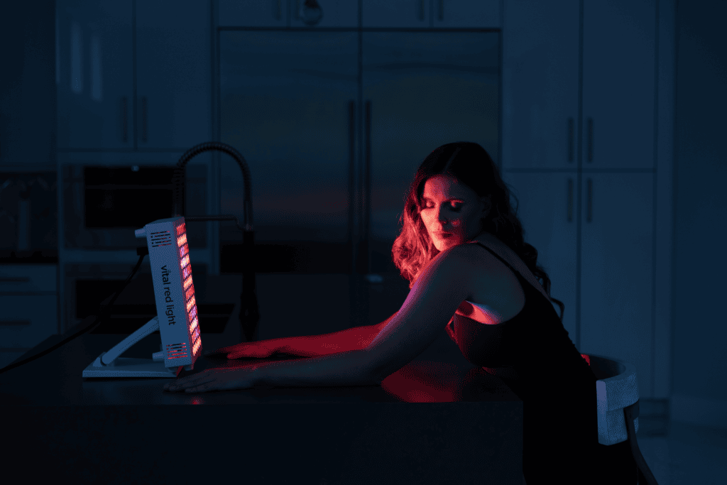 A woman sits at a kitchen counter, leaning toward the Vital Pro 2.0 red light therapy panel that illuminates her face and arms.